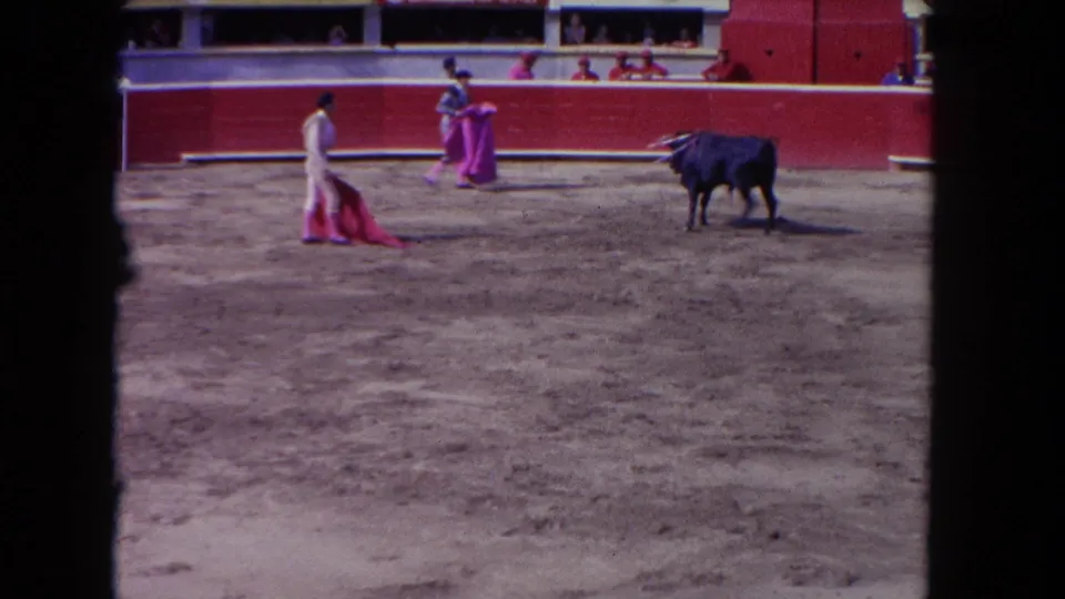 8mm home-movie frame circa 1962 showing two arena performers — one in white, one in pink — facing a stationary black bull, with spectators in tiered seating behind the barrier.