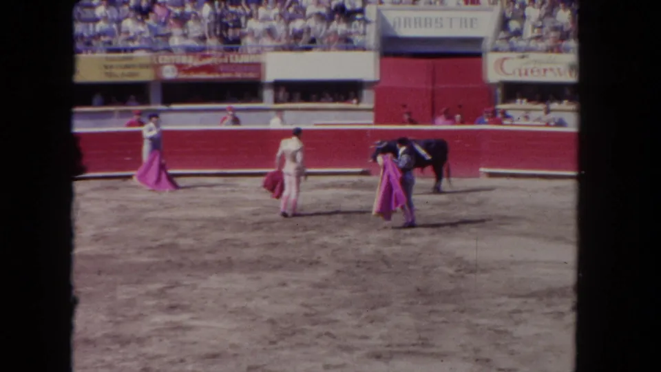 Multiple toreros and a dark bull in action in a sunlit arena, circa 1962; Cuervo advertising and ARRASTRE banner visible on the stands walls.
