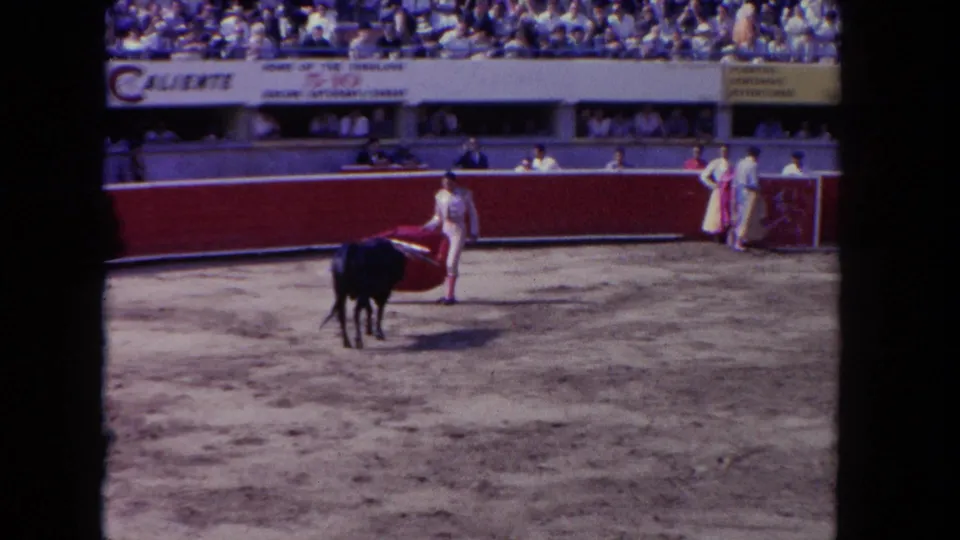 1962 8mm color frame of a matador in traje de luces holding a magenta cape before a black bull; Caliente venue banners visible in the background stands.