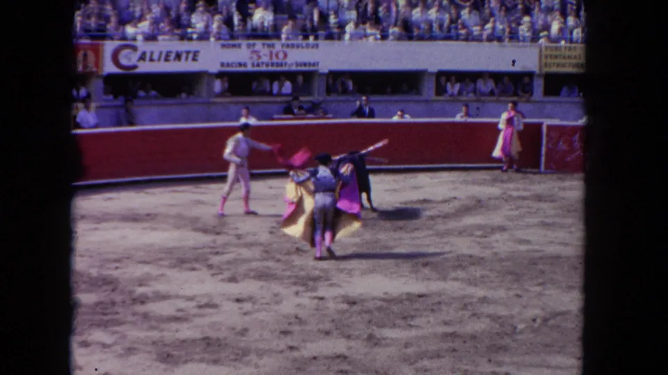 8mm color frame c. 1962 showing two toreros working a black bull near the barrera; Caliente venue banners and a dense grandstand crowd visible above.