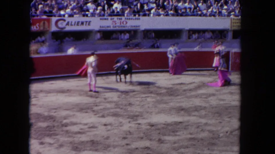 1962 8mm frame of a bullfight, likely in Tijuana, Mexico; matadors with magenta capes confront a bull while spectators fill the stands behind a red barrera.