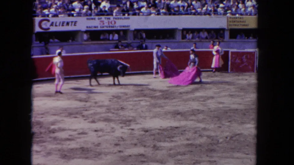 1962 8mm frame: charging bull and matador with magenta cape in foreground; multiple performers in ring; Caliente advertising banner prominent above packed stands, likely Tijuana.