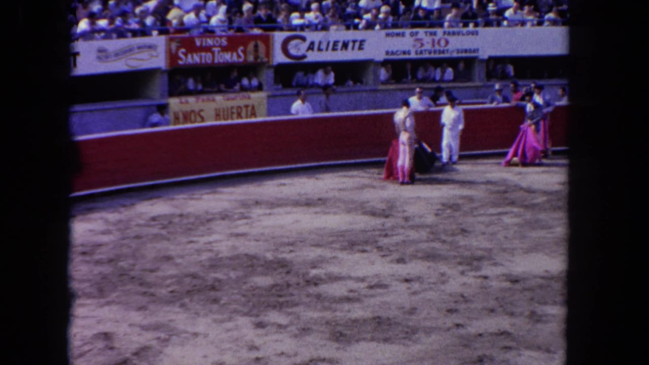 Wide shot of the same bullfighting arena; toreros in colorful costumes repositioned near arena center, crowd visible in grandstands above.