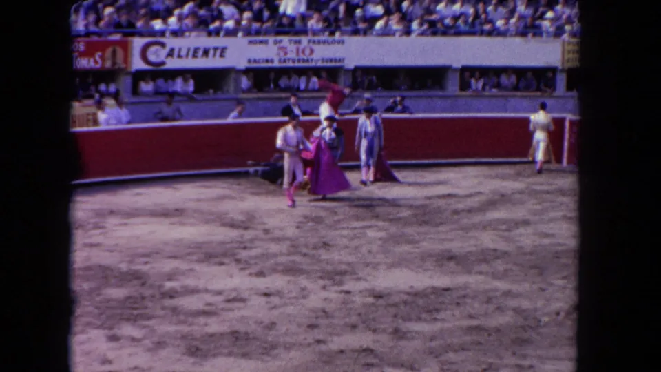 8mm home-movie frame circa 1962 showing two matadors in corrida ring with Caliente racing banner and crowded grandstand.