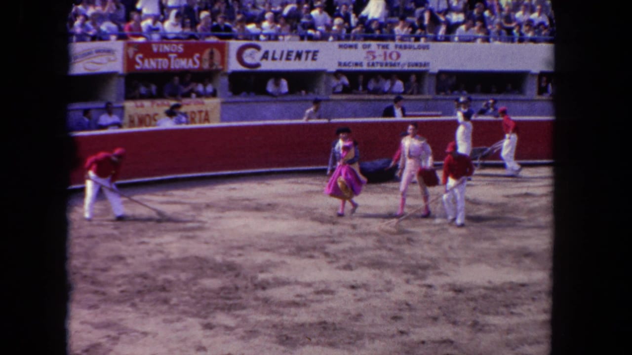 Bullfighting arena with costumed matadors and white-uniformed workers on sand, Caliente and Vinos Santo Tomas banners above packed stands.