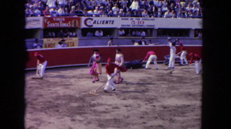 Arena cleanup crew raking sand circa 1962 as matadors in pink and magenta costumes walk nearby; Caliente and Vinos Santo Tomas banners span the stands wall.
