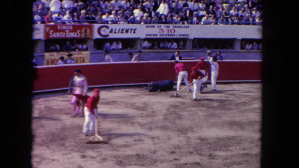 Post-fight arena raking scene circa 1962, workers in white and a costumed figure on sand; Caliente and Vinos Santo Tomas banners above the red barrera wall.