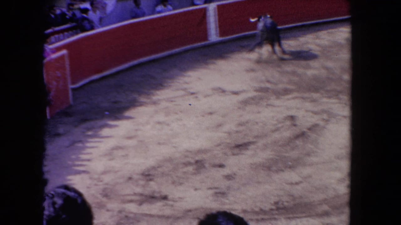Overhead 8mm view of a bullfighting arena; a lone figure stands in the sandy ring center while crowds fill the upper stands.