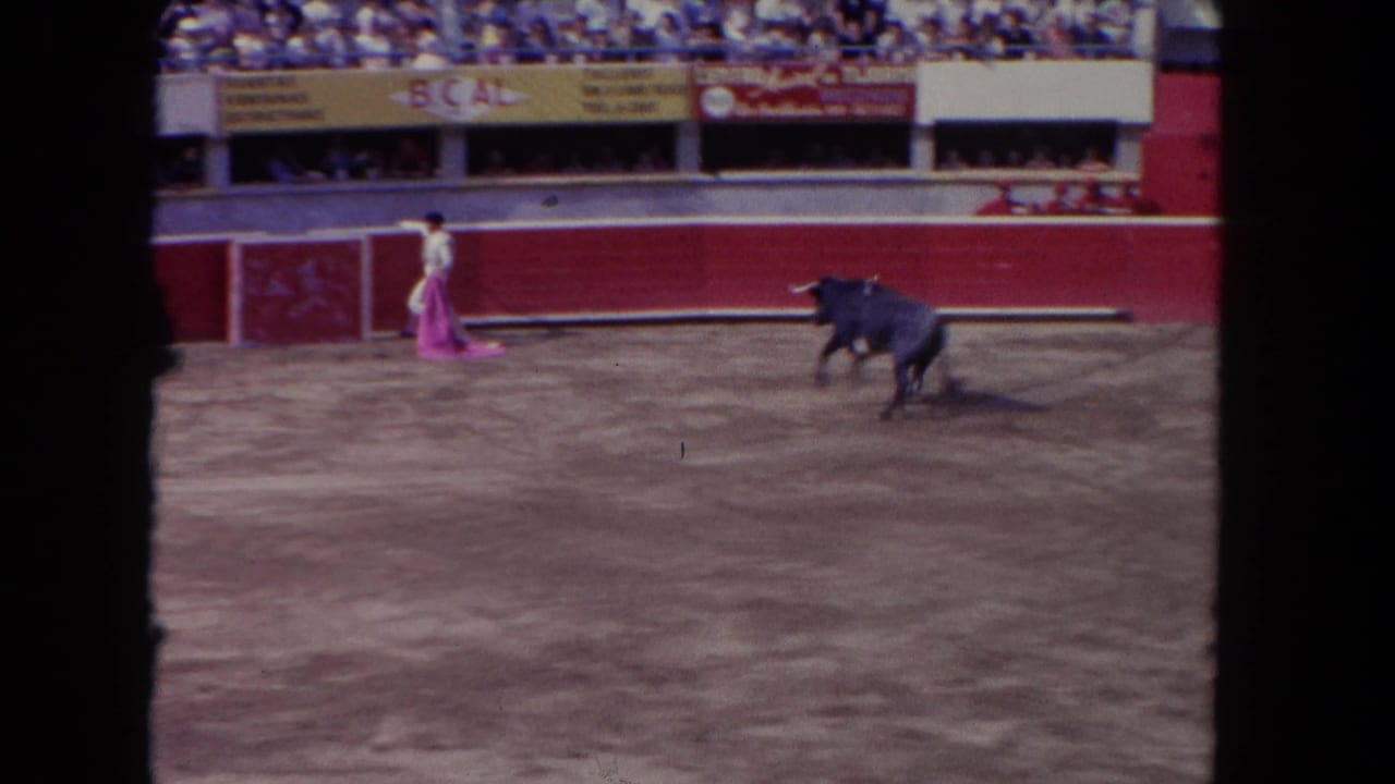 8mm frame circa 1962 showing matador in pink facing a charging dark bull across the sandy arena floor, spectators in distant stands.