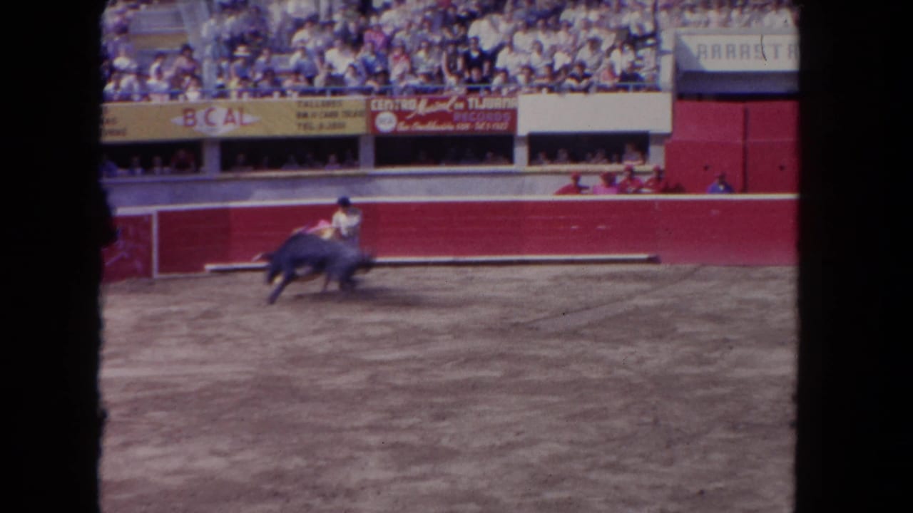 Wide arena shot c.1962, possibly Tijuana, Mexico; matador and bull in the ring, 'B.CAL' and 'CENTRO' banners on red barriers, spectators filling the stands.