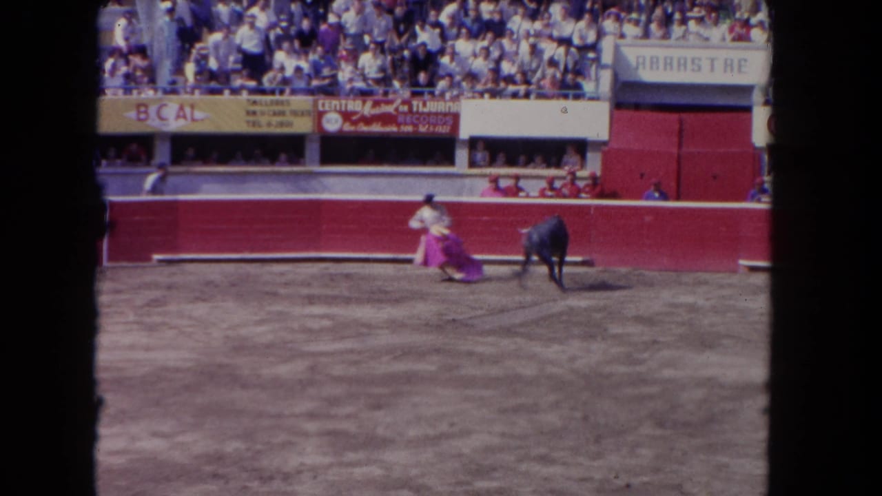 8mm home movie, c. 1962 — wide shot of corrida arena; matador executing a cape pass with a dark bull, advertising banners including 'BCAL' legible on the red barrera.