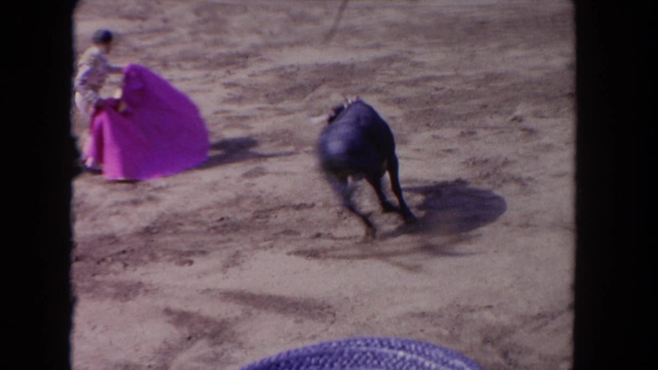 1960s 8mm frame of a matador with a pink cape facing a bull in a sandy bullring, seen from an elevated spectator angle.