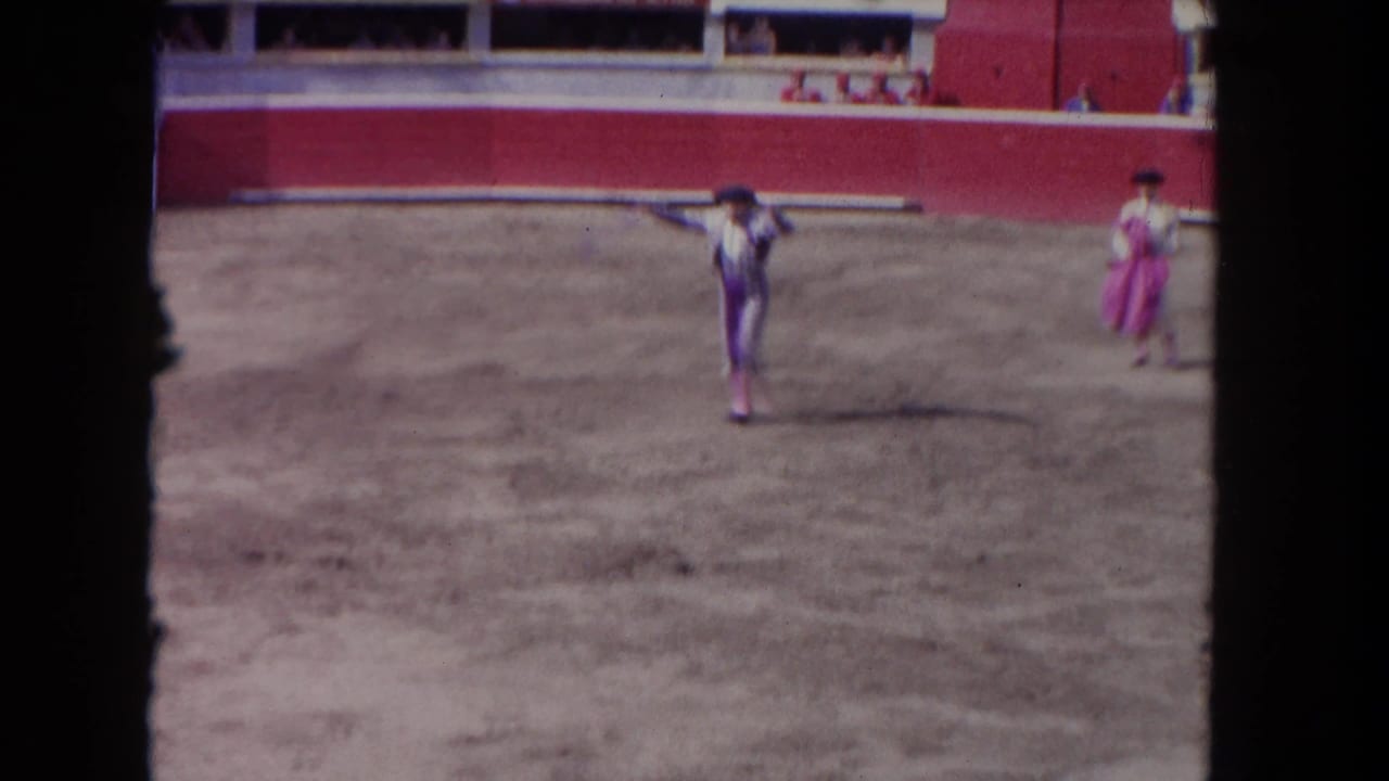 Matador in a sandy arena with a pink-caped figure nearby and red arena wall behind.