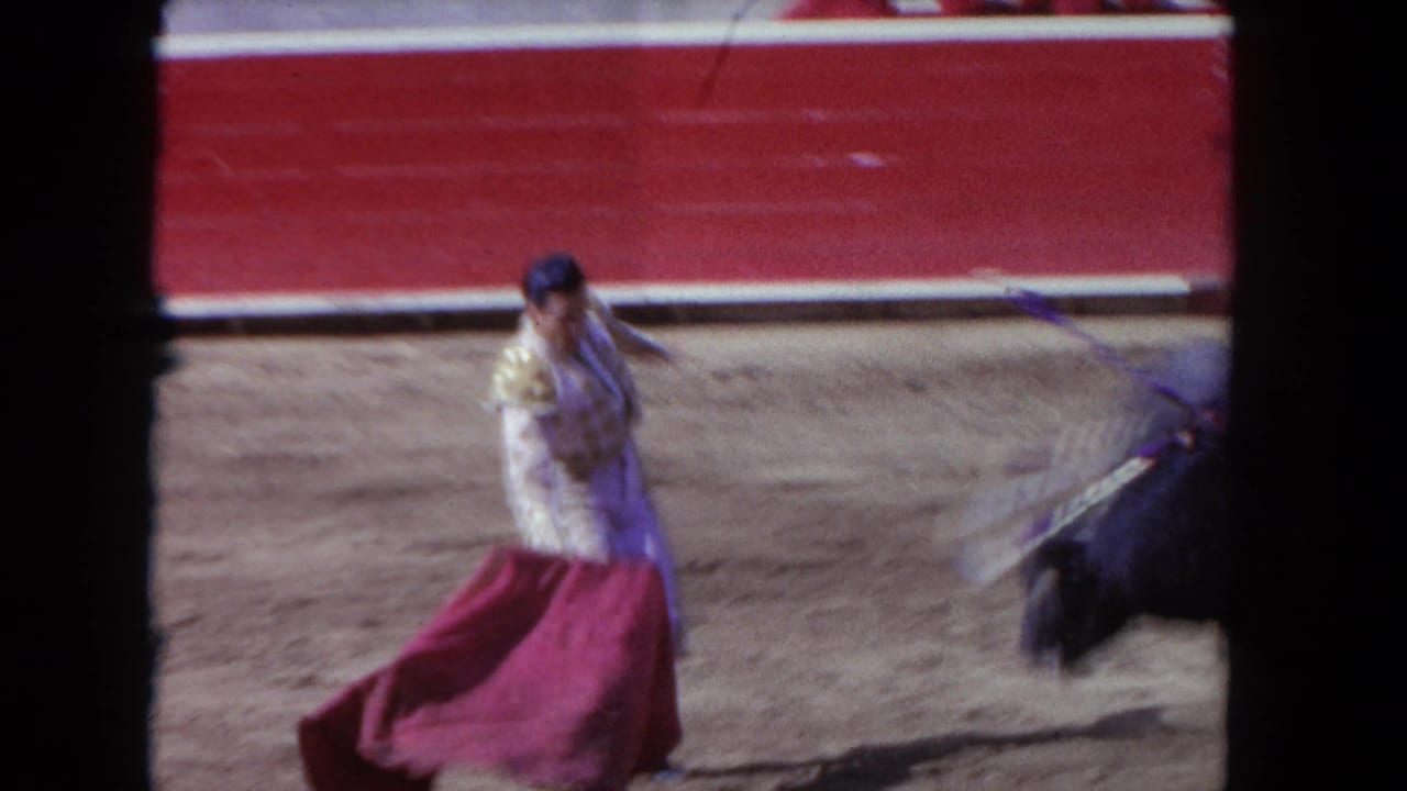 Matador in ornate traje de luces extends a red muleta toward a charging dark bull; red barrera wall and sand floor visible; 8mm home-movie footage, circa 1962.