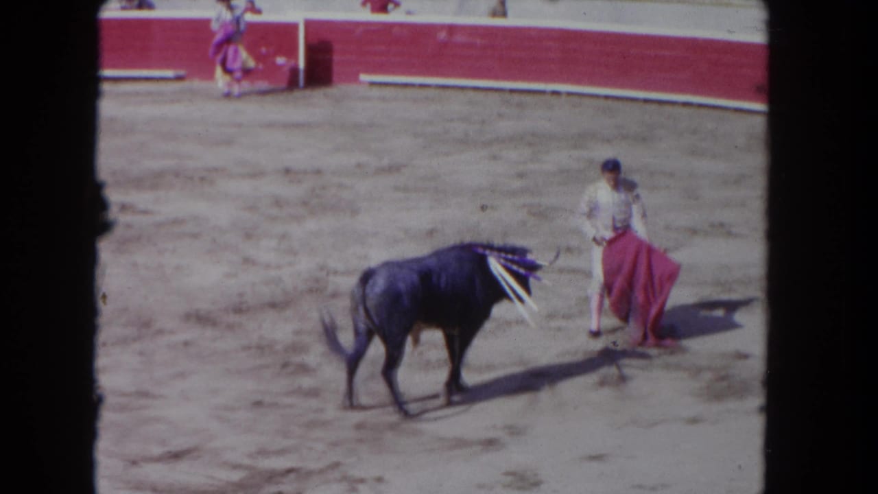 1962 8mm home-movie frame of a matador working a red muleta pass against a dark bull with banderillas planted in its shoulders, filmed from the stands.