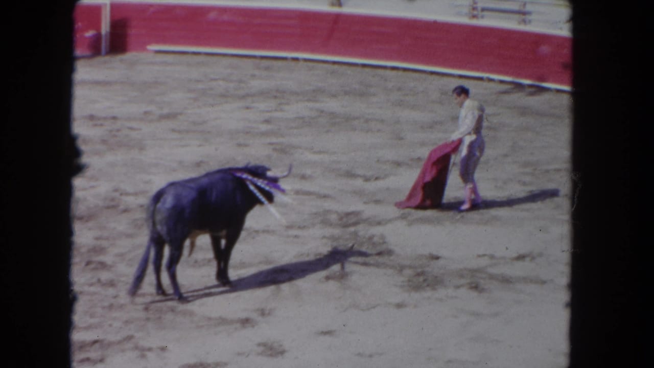 1962 8mm home-movie frame showing matador and bull at mid-pass pause; banderillas visible in bull's shoulders in a corrida arena.