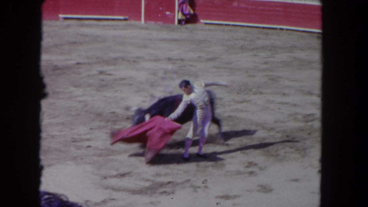 Matador in pale suit bending low, extending a pink-red cape close to a large dark bull in a sandy arena.