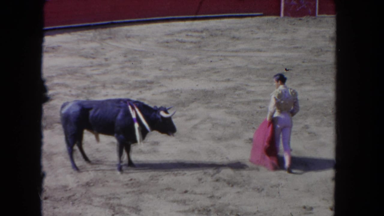 Matador in embroidered light costume holds pink muleta facing a dark bull with bandilleras, in a bullfight arena circa 1962 per metadata.
