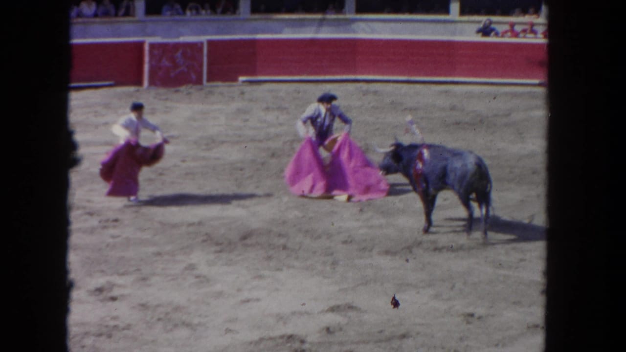 Wide 8mm view of bullfighting action with two matadors, a dark bull, a red-walled barrera, and spectators in the upper stands, consistent with early 1960s footage.
