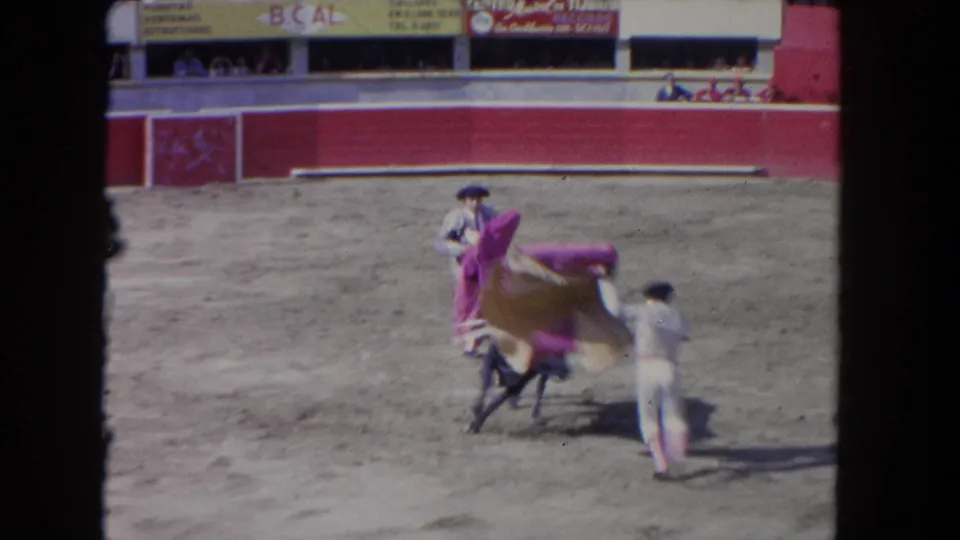 8mm close-up of a cape pass during a bullfighting event; advertising banners are visible on the red barrier wall, consistent with early 1960s arena footage.