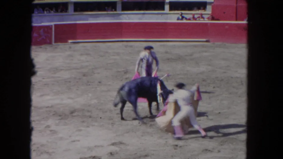8mm frame of a matador in very close proximity to a charging dark bull in the sand arena, with spectators visible in the stands beyond the red barrera.