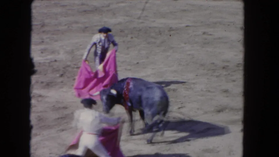 Close bullfight action circa 1962; matador in ornate blue-silver traje de luces holds pink capote as a dark bull charges; redness visible on bull's neck area, second matador in foreground.