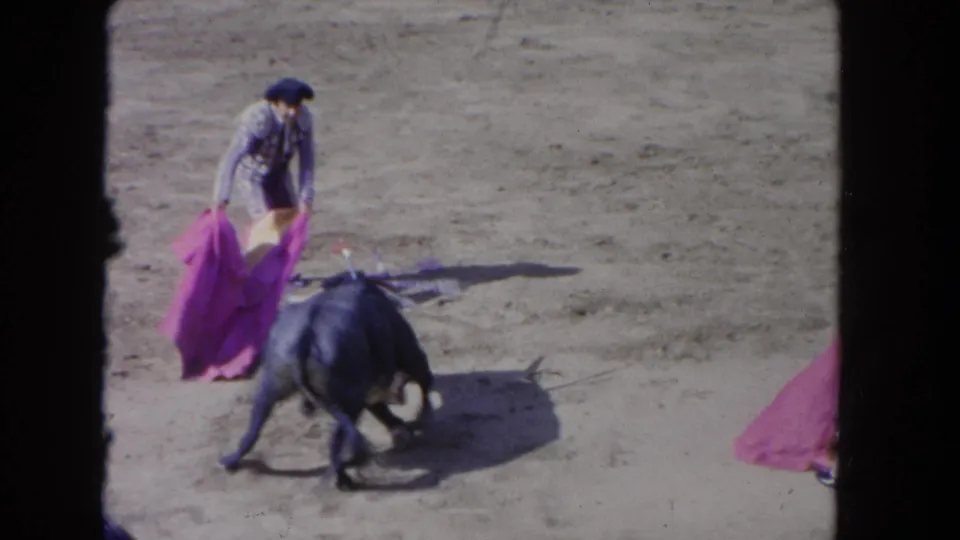 Bullfight arena circa 1962; matador in traje de luces directs a charging dark bull using a large pink capote, partial second figure visible at frame edge.