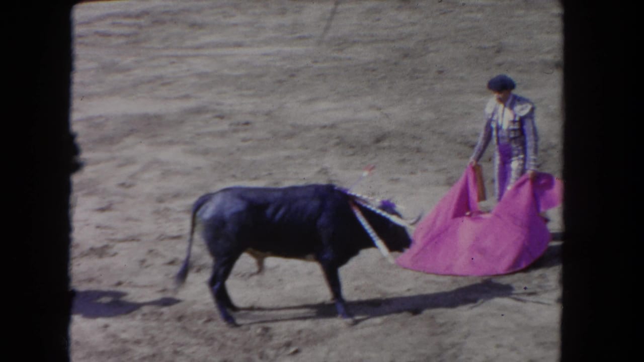 8mm footage circa 1962: matador in silver-purple traje de luces extends magenta capote toward a charging bull in a dusty outdoor arena