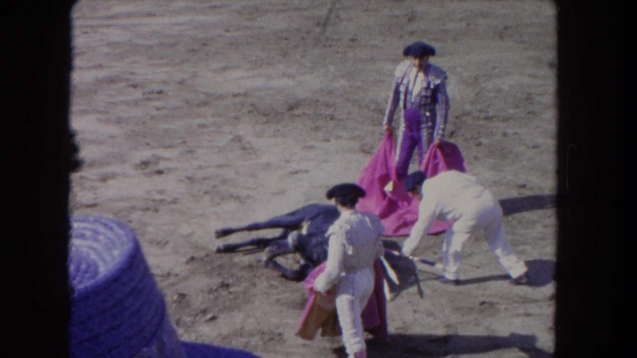 1962 8mm home-movie frame of a bullfighting finale: matador in purple traje de luces, fallen bull, and peon with magenta cape in a sandy arena, likely Los Angeles.