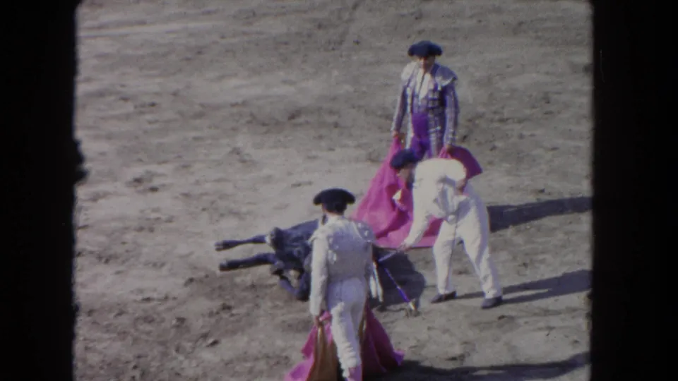 8mm home-movie frame circa 1962 showing a matador in purple traje de luces with cape, a white-clad peon, and a fallen bull on a sandy arena floor.