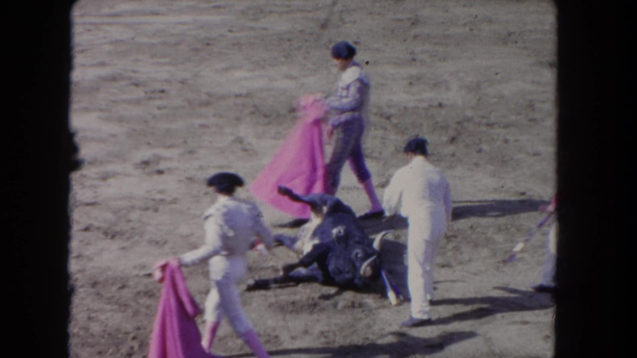 Bull lying on sandy arena floor; matador in white ornate suit holds pink cape; second figure in white stands nearby.