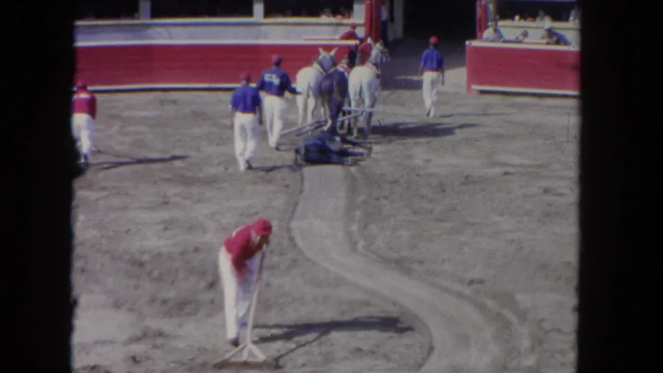 8mm color film circa 1962: workers in red-and-white uniforms rake a dirt arena floor at an outdoor venue, with a white horse and blue-uniformed handlers visible behind.