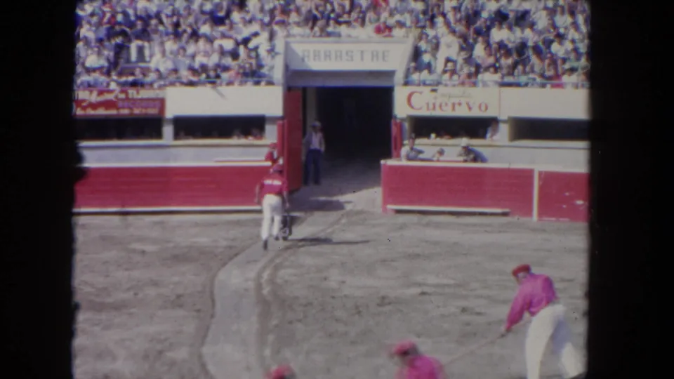 8mm color film circa 1962: arena floor workers in red uniforms, a white horse with handlers near red barriers, and full tiered stands with spectators; Cuervo brand sign visible on facade.
