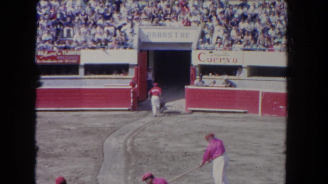 Arena worker in pink-and-white costume sweeps the corrida floor, ca. 1962; red-and-white painted wooden barriers with José Cuervo advertising and a full grandstand crowd visible.