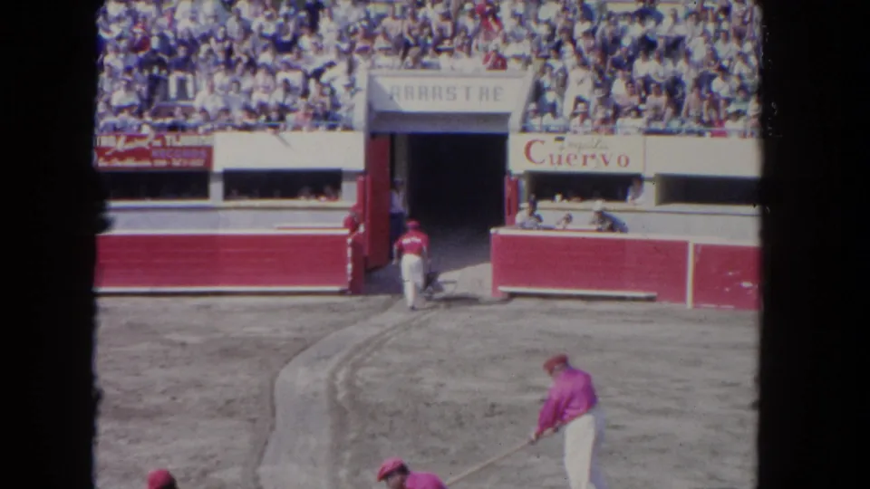 Arena worker in pink-and-white costume sweeps the corrida floor, ca. 1962; red-and-white painted wooden barriers with José Cuervo advertising and a full grandstand crowd visible.