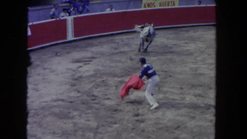 8mm wide shot of bullring: matador in blue suit of lights holding red muleta cape, white horse in the arena, circular barrera with Hnos Huerta banner, tiered stands with spectators.