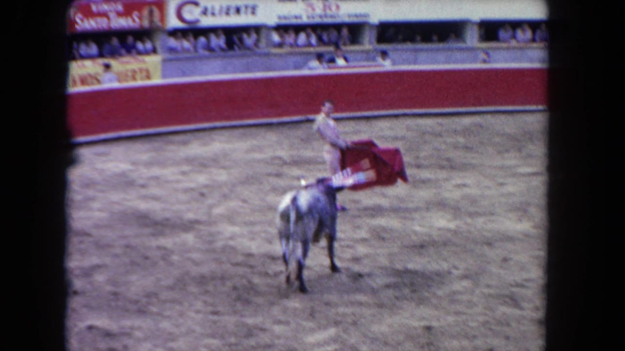 Matador in light corrida suit with pink stockings extends a red muleta cape toward a dark bull in a Caliente-branded bullfighting arena, circa 1962.