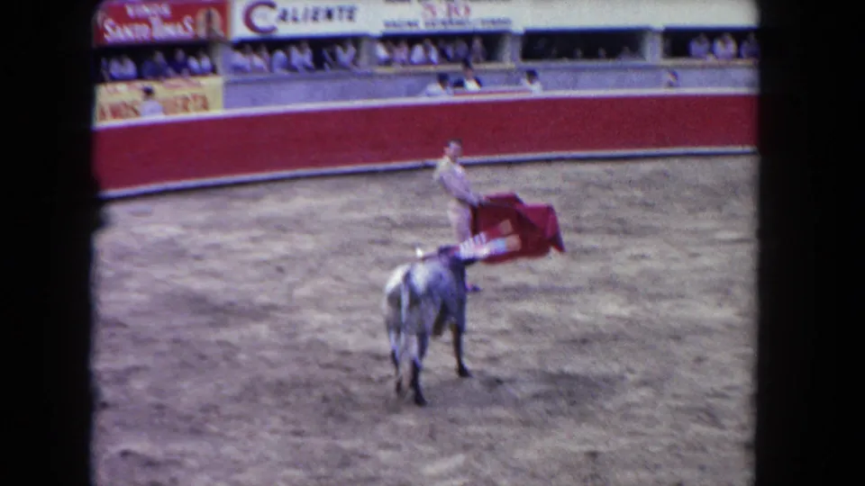 Matador in light corrida suit with pink stockings extends a red muleta cape toward a dark bull in a Caliente-branded bullfighting arena, circa 1962.