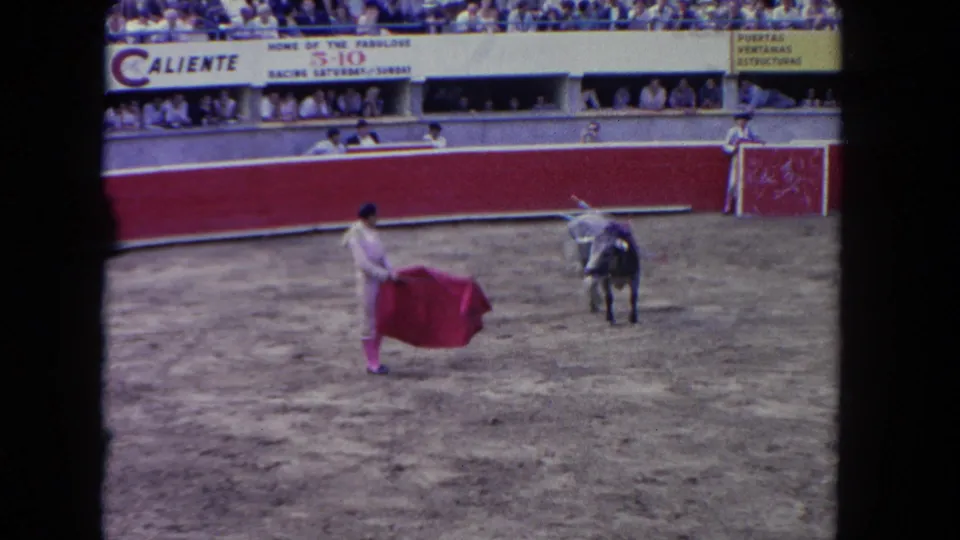 Wide view of a Caliente-branded bullfighting arena showing a matador in pink suit with lowered muleta, a bull with banderillas at distance, and full grandstands, circa 1962.