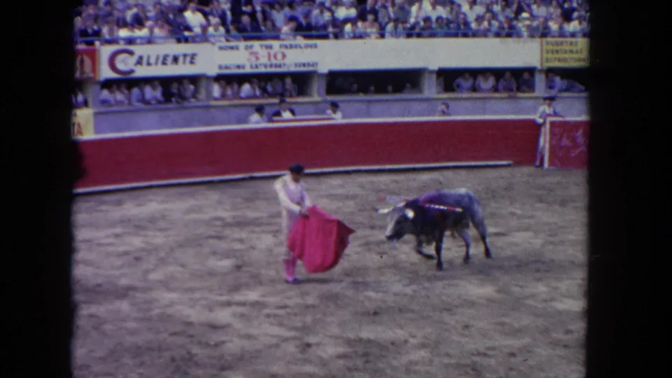 Matador in light corrida suit works a red muleta at close range to a dark bull with banderillas in a Caliente-branded arena with packed grandstands, circa 1962.