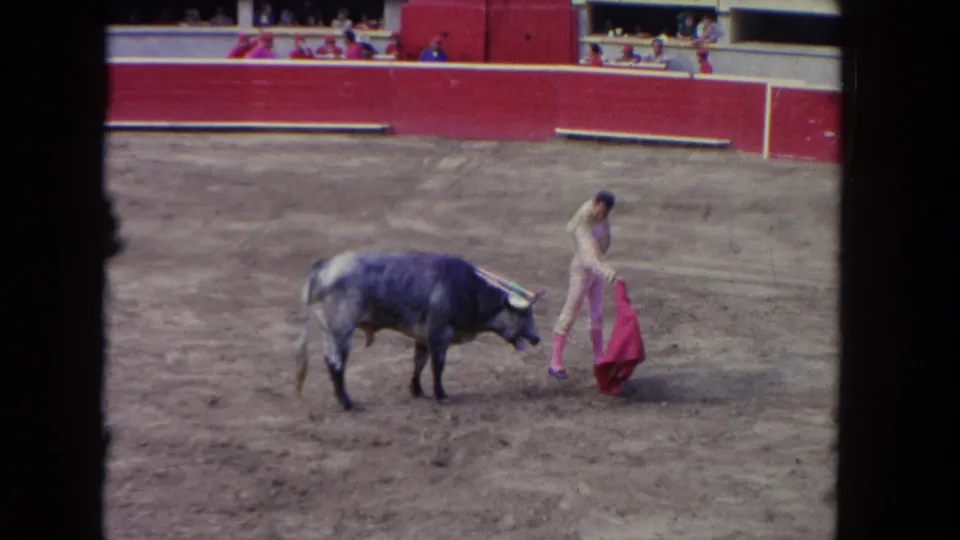Wide 8mm home-movie frame circa 1962: matador facing a bull in the center of a bullfighting ring; red wooden barrera and spectator stands occupy the background.