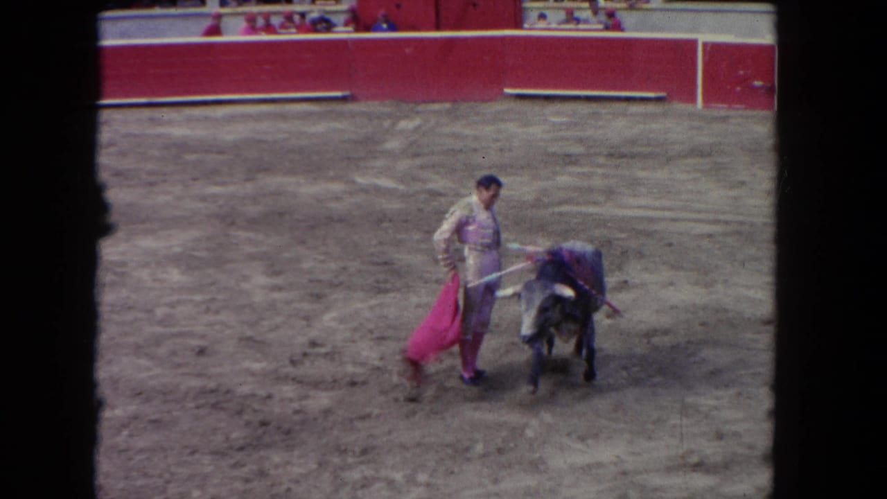 Wide shot of a matador in a cream traje de luces passing a dark bull in a red-barrered arena; 8mm footage circa 1962.