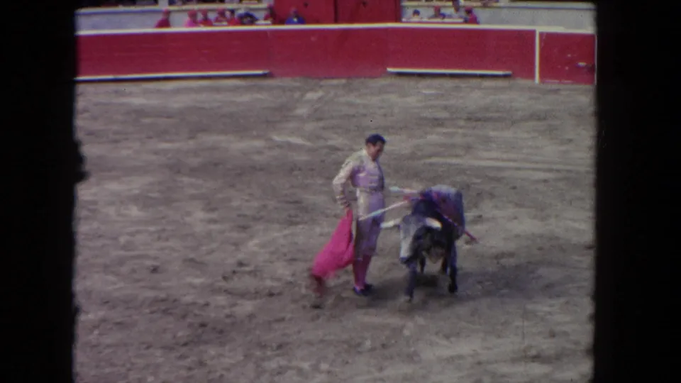Wide shot of a matador in a cream traje de luces passing a dark bull in a red-barrered arena; 8mm footage circa 1962.