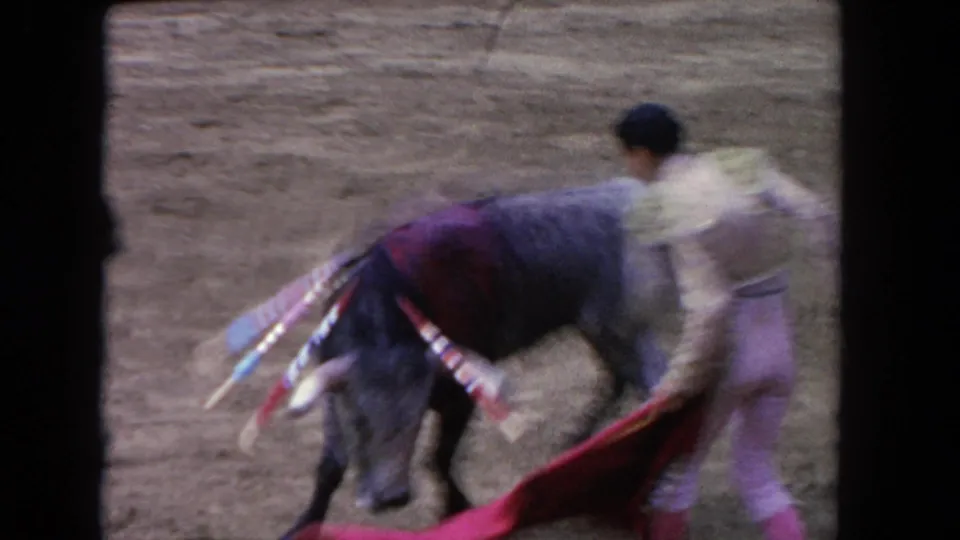 Motion-blurred 8mm frame capturing a matador executing a muleta pass as the bull charges through in a corrida arena, circa 1962.