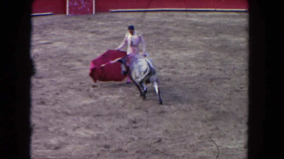 Wide spectator-distance view of matador extending red cape toward charging bull in a sandy bullfighting arena, circa 1962