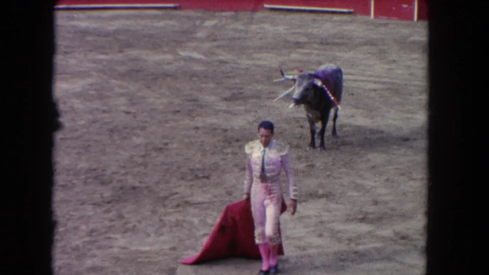 1962 8mm: matador in traje de luces faces camera with red muleta at ground level; dark bull stands close behind in sandy arena