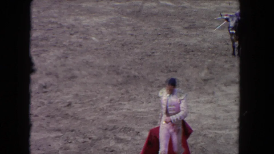 1962 8mm: matador pauses with red muleta in open arena; bull visible at frame right; shot from elevated spectator stands