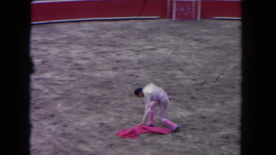 8mm frame circa 1962 showing a lone matador in a light suit bending to retrieve a pink muleta on the cleared arena floor after the bull has been removed.