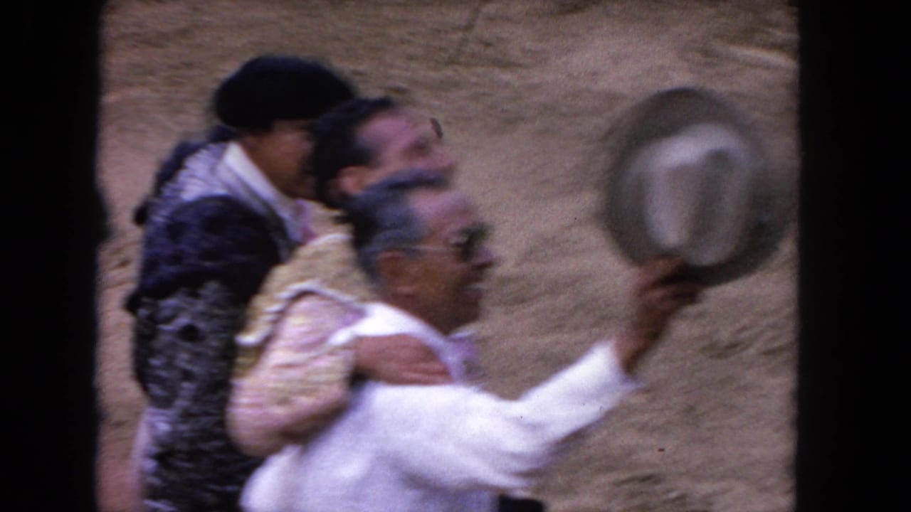 Bullfighter and older companion embrace in the arena, the companion's arm raised with a hat blurred in motion overhead.