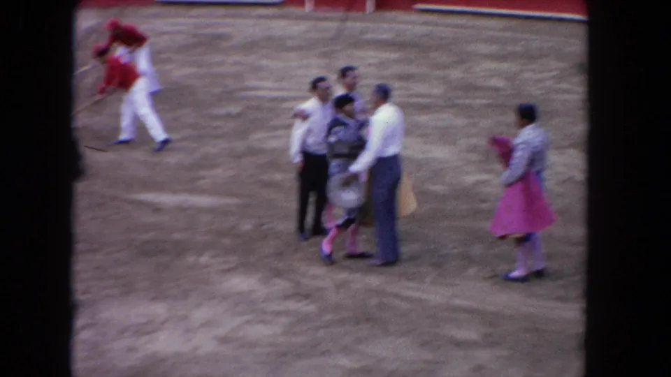 Matadors in pink costumes and civilian-dressed men gather on arena sand near a red barrier wall, Los Angeles circa 1962.